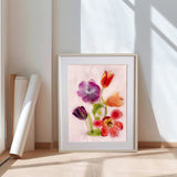 A framed watercolor floral art print leaning against a white wall on a light wood floor, with rolled papers beside it. The artwork shows tulips and other flowers in pink, purple, orange, red, and green tones on a pale pink background.